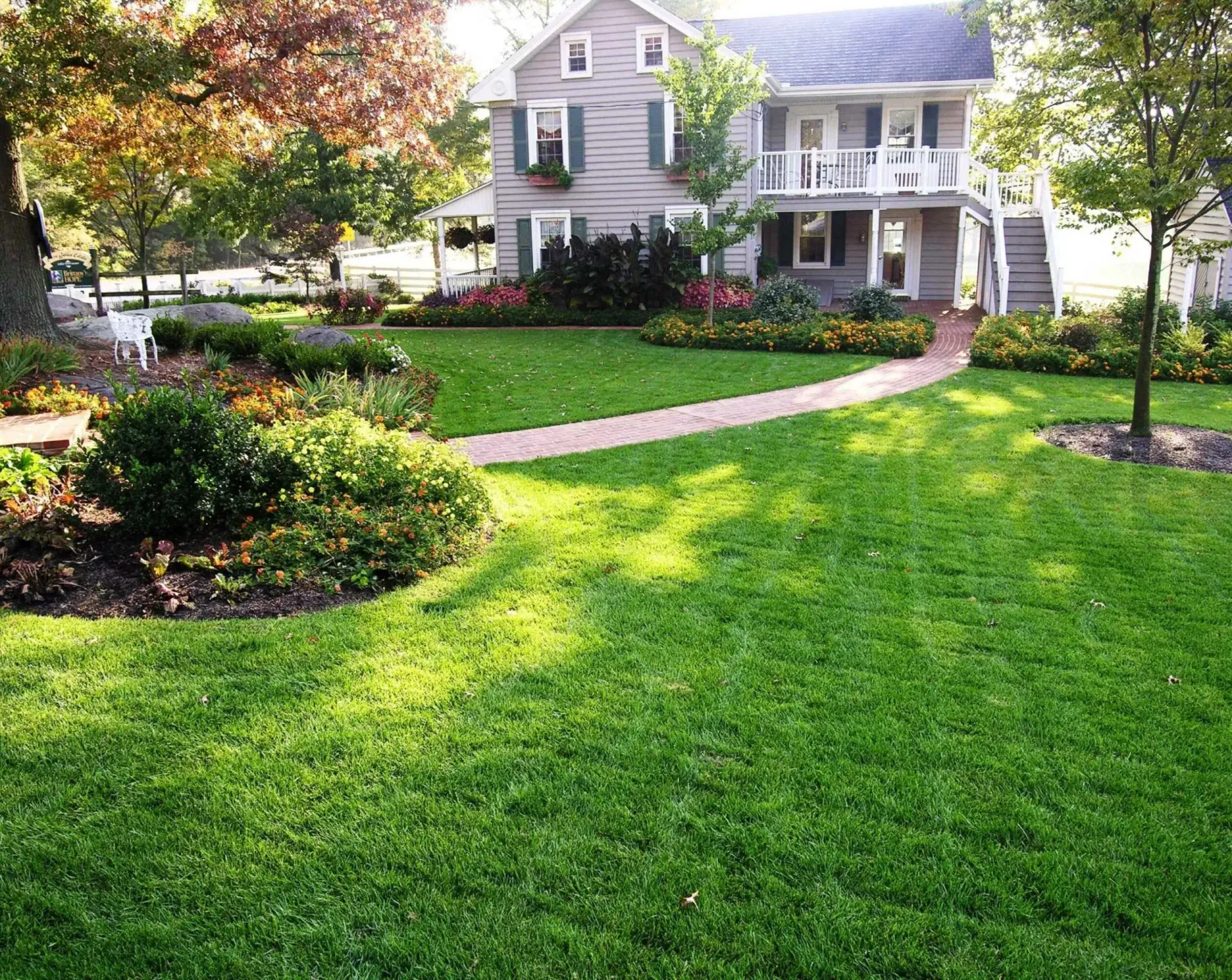Well-manicured lawn and brick pathway leading to a two-story house with shutters, surrounded by trees and flower beds.