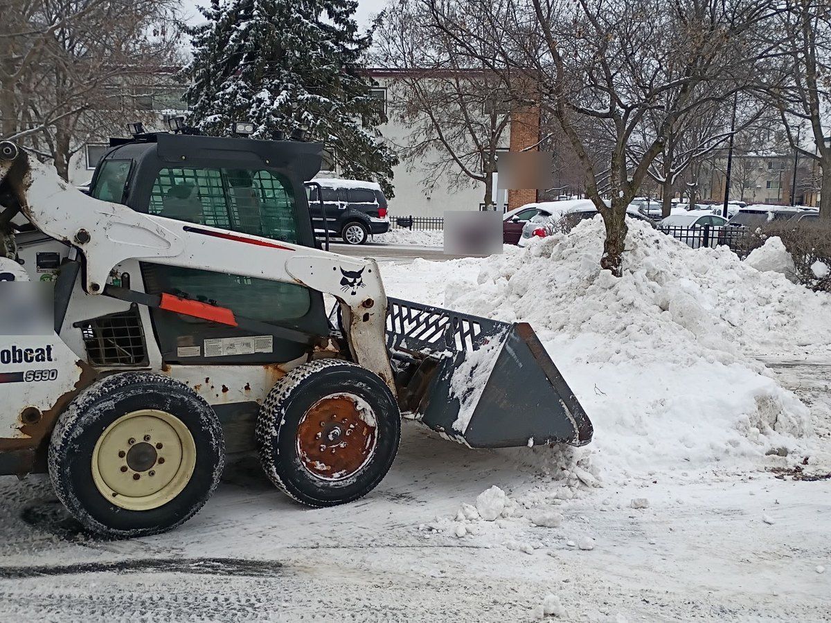 Skid loader working near a snow pile with parked cars in the lot behind it