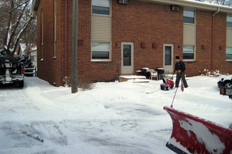 Person using a snowblower to clear snow from a driveway in front of a brick apartment building.