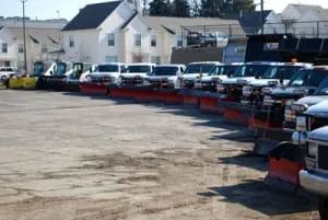 Line of snowplow trucks parked on a lot in front of houses.