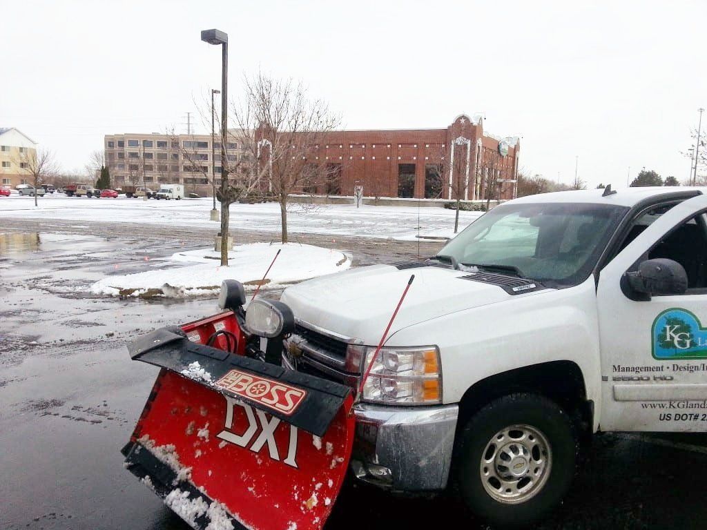 A white pickup truck with a red Boss snow plow attached to the front sits in a wet, snowy parking lot.