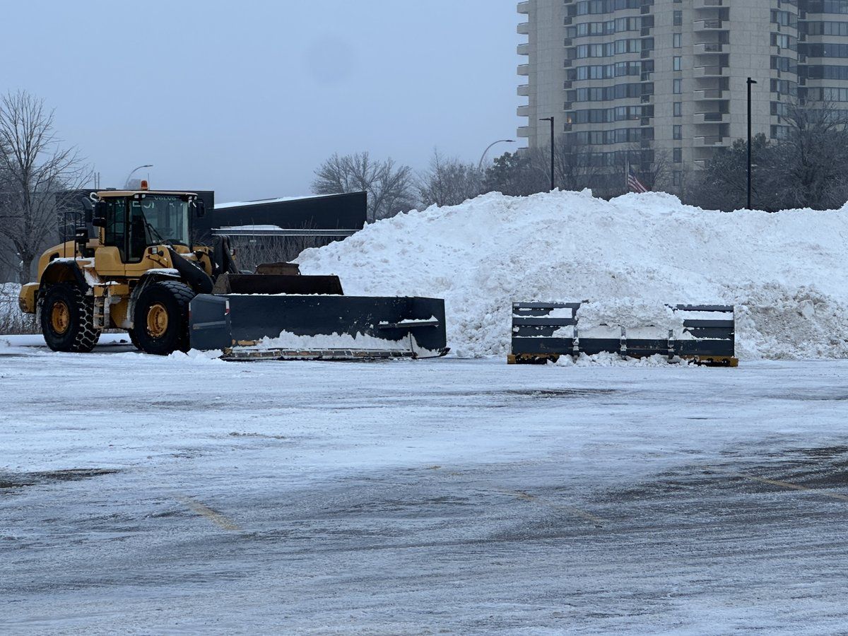 Wheel loader with a pusher box beside a large pile of stacked snow in a parking lot