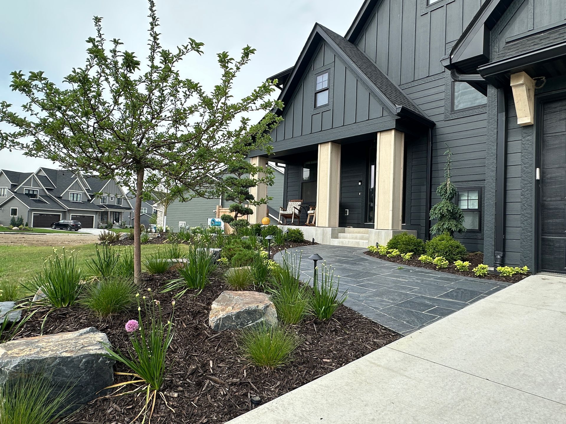Front yard with black flagstone walkway, mounded plant beds, gray boulder outcroppings, and a tree in Plymouth MN