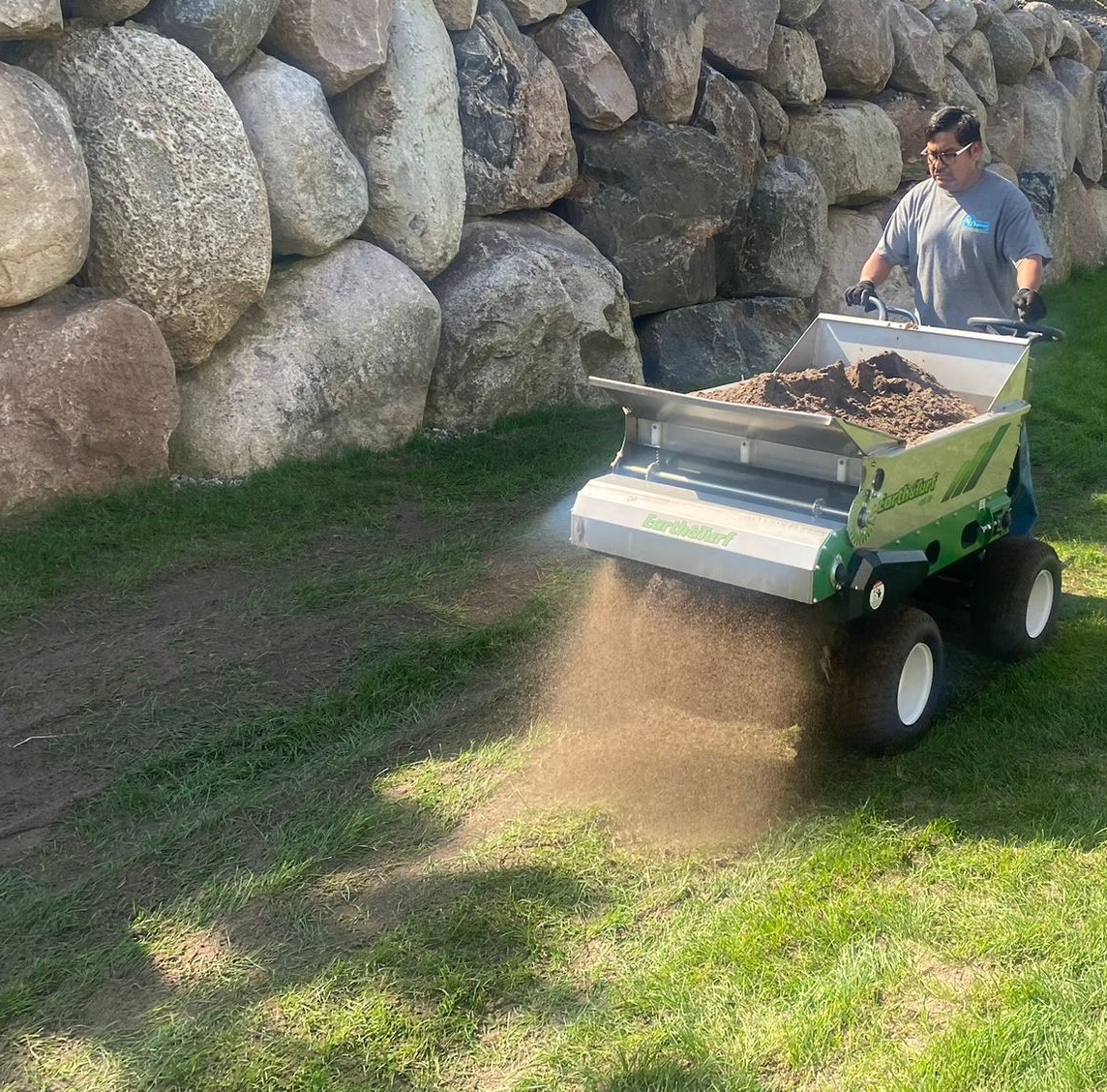A person pushes a green topdresser machine, spreading soil across a grassy lawn in front of a large stone retaining wall.