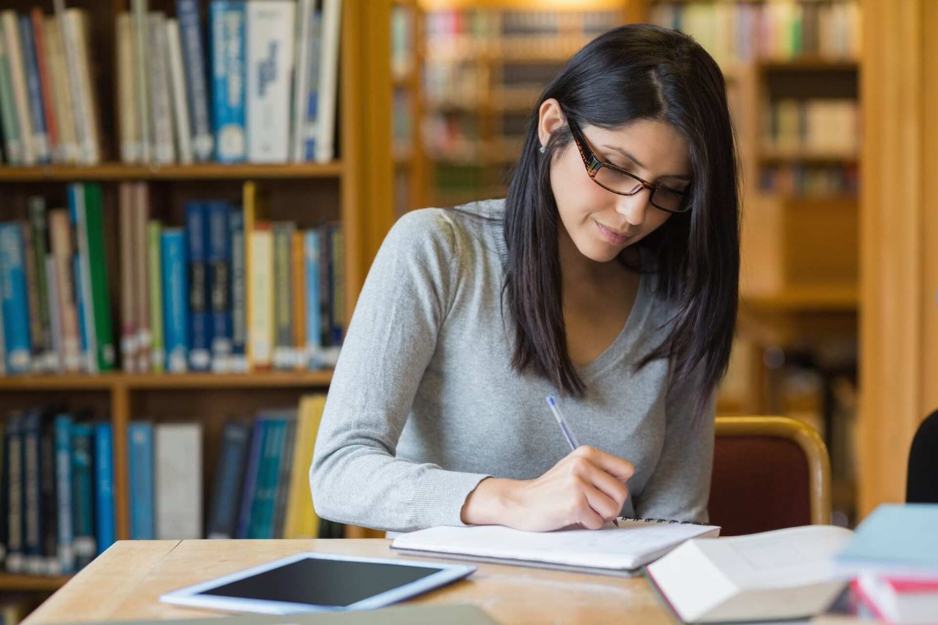 Woman with glasses writing in a notebook at a table in a library. A tablet and books are present.