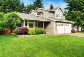 Two-story house with beige siding, white garage doors, green lawn, and vibrant landscaping.