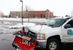 White pickup truck with snowplow parked in a snowy area, a building is in the background.