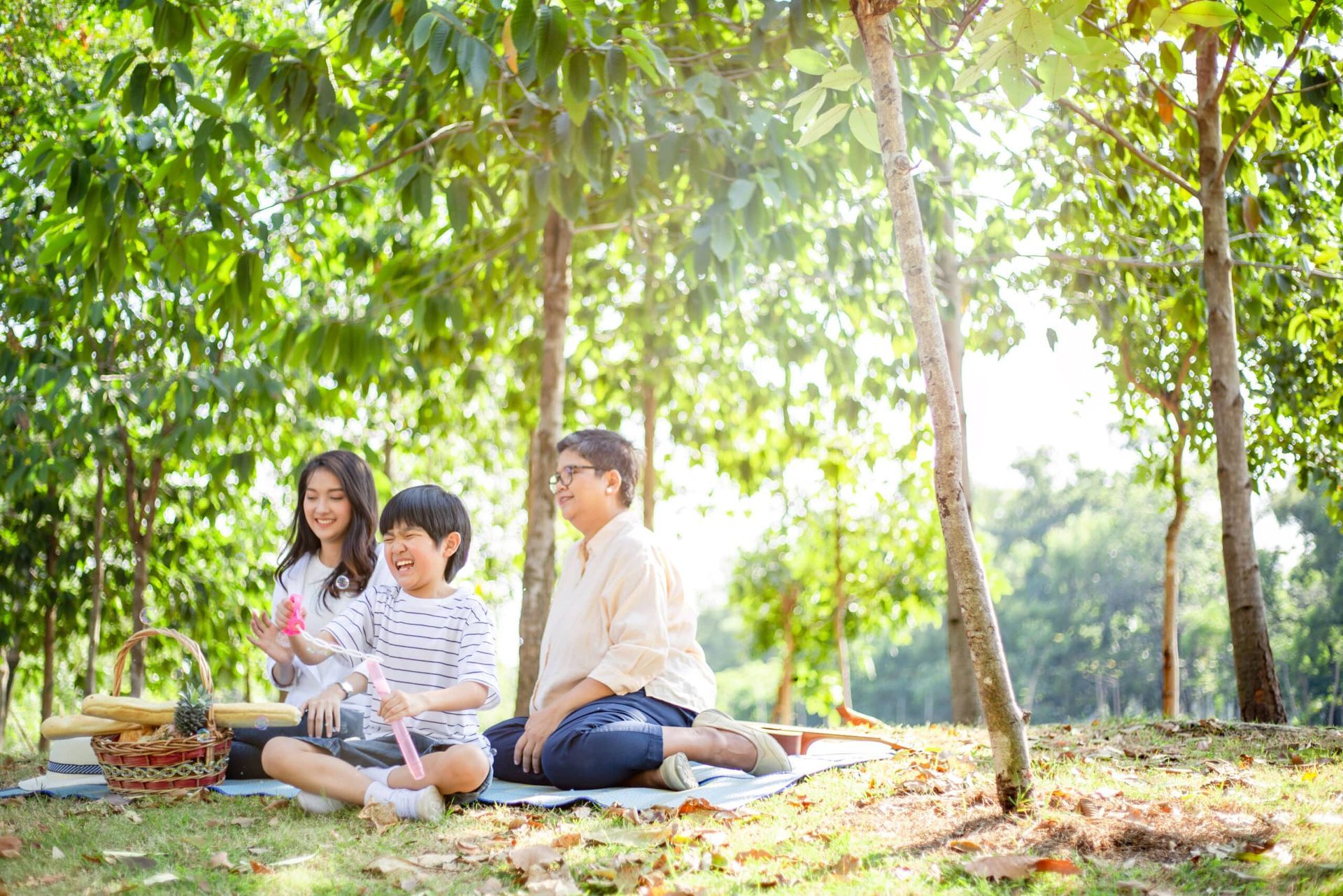 Three people picnic on a blanket under trees in a sunlit park, with baskets and food nearby.