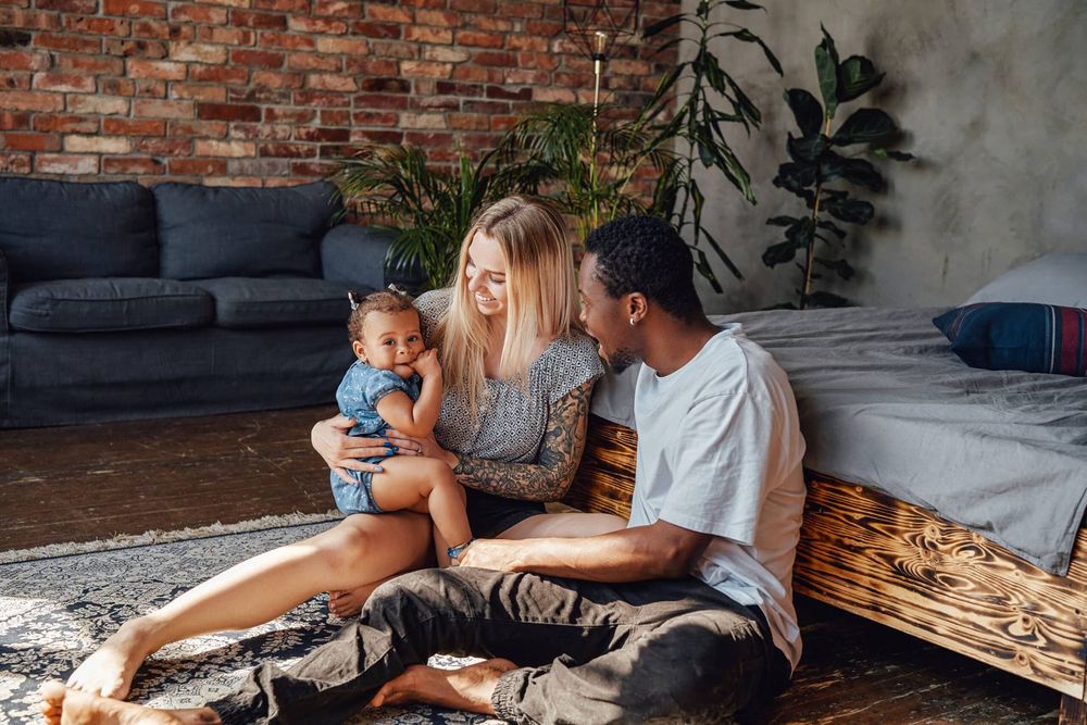 Family sitting on a rug in a cozy living room, with a baby between two adults near a couch and bed.