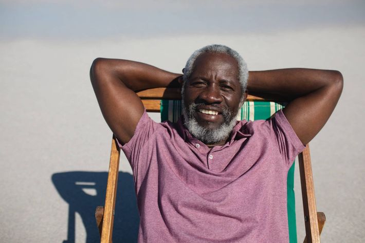 Smiling man in a pink polo reclines with hands behind his head on a striped chair outdoors.