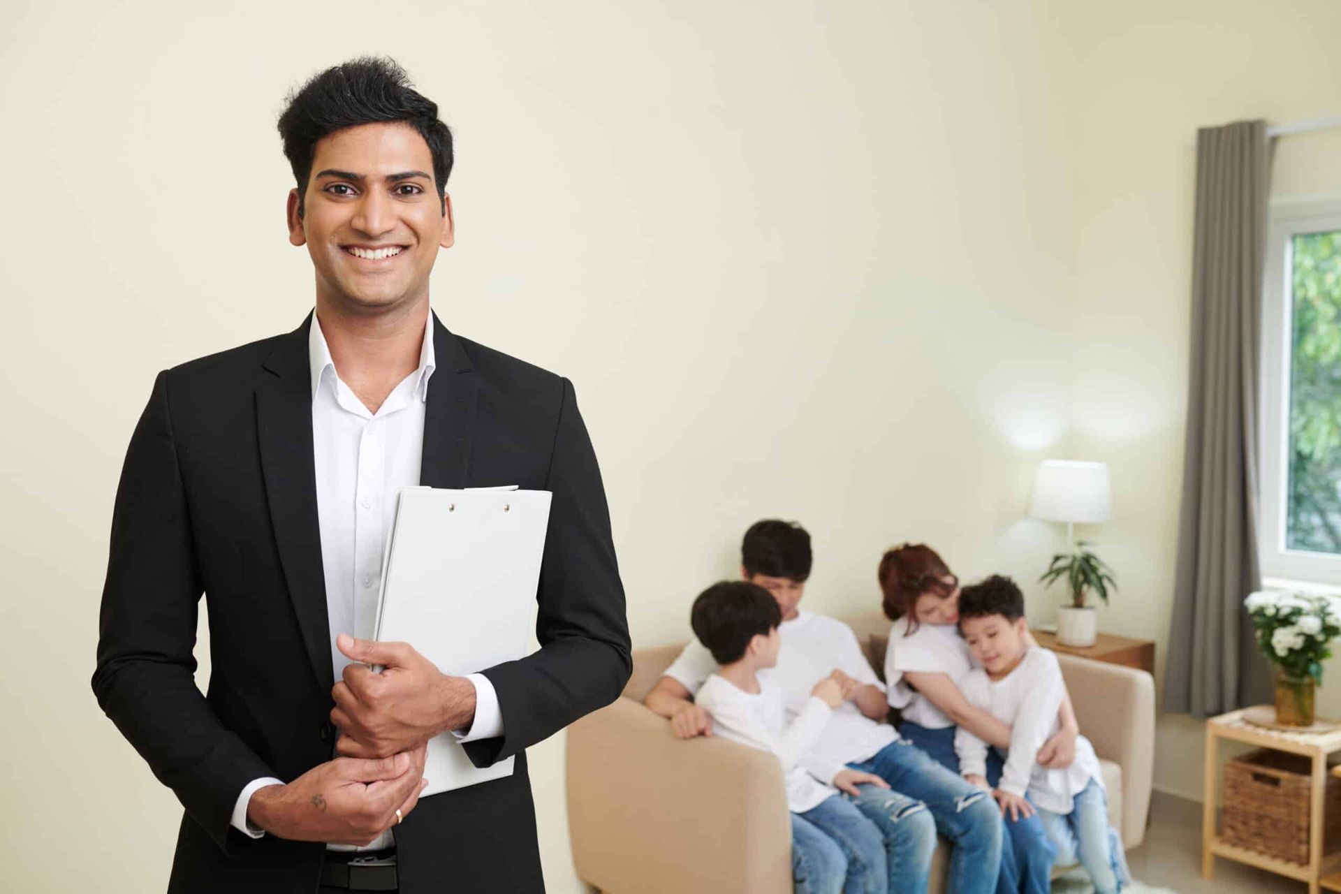 Man in black blazer holding papers, standing beside a seated group in a bright living room