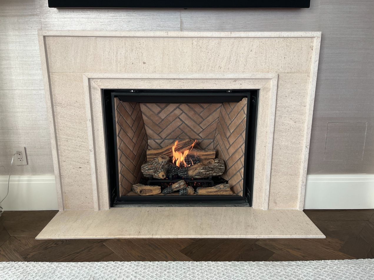 Lit gas fireplace with beige stone surround and herringbone firebox in a neutral living room