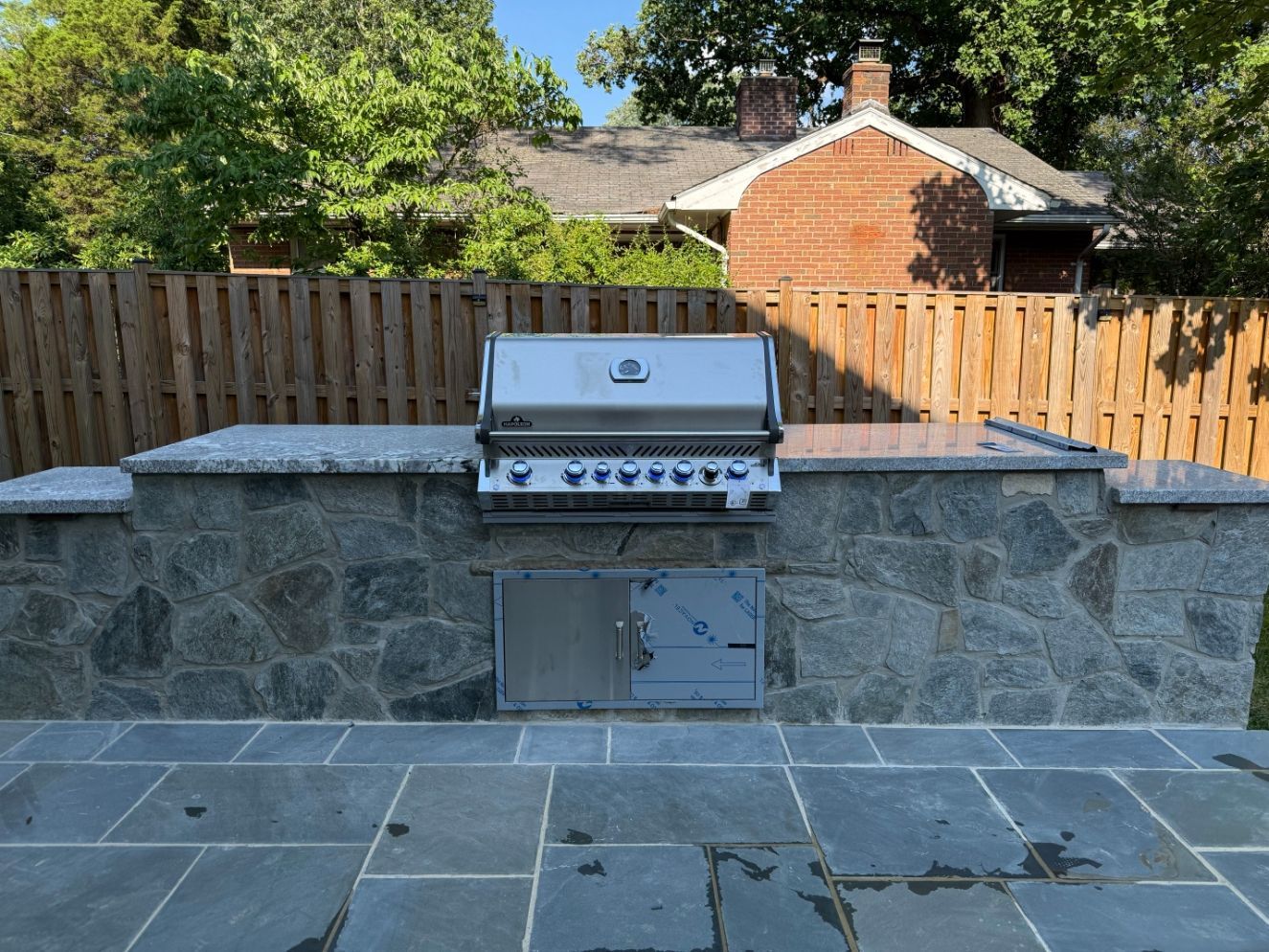 Outdoor stone patio with built-in stainless grill and low stone wall in a fenced backyard