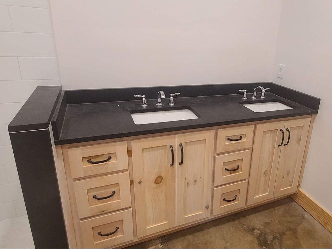 A bathroom vanity with two sinks. It has light wood cabinets and a black countertop.