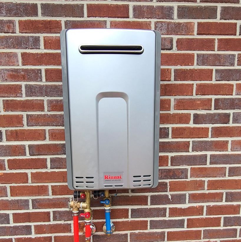 A silver tankless water heater on a brick wall with red and blue pipes.