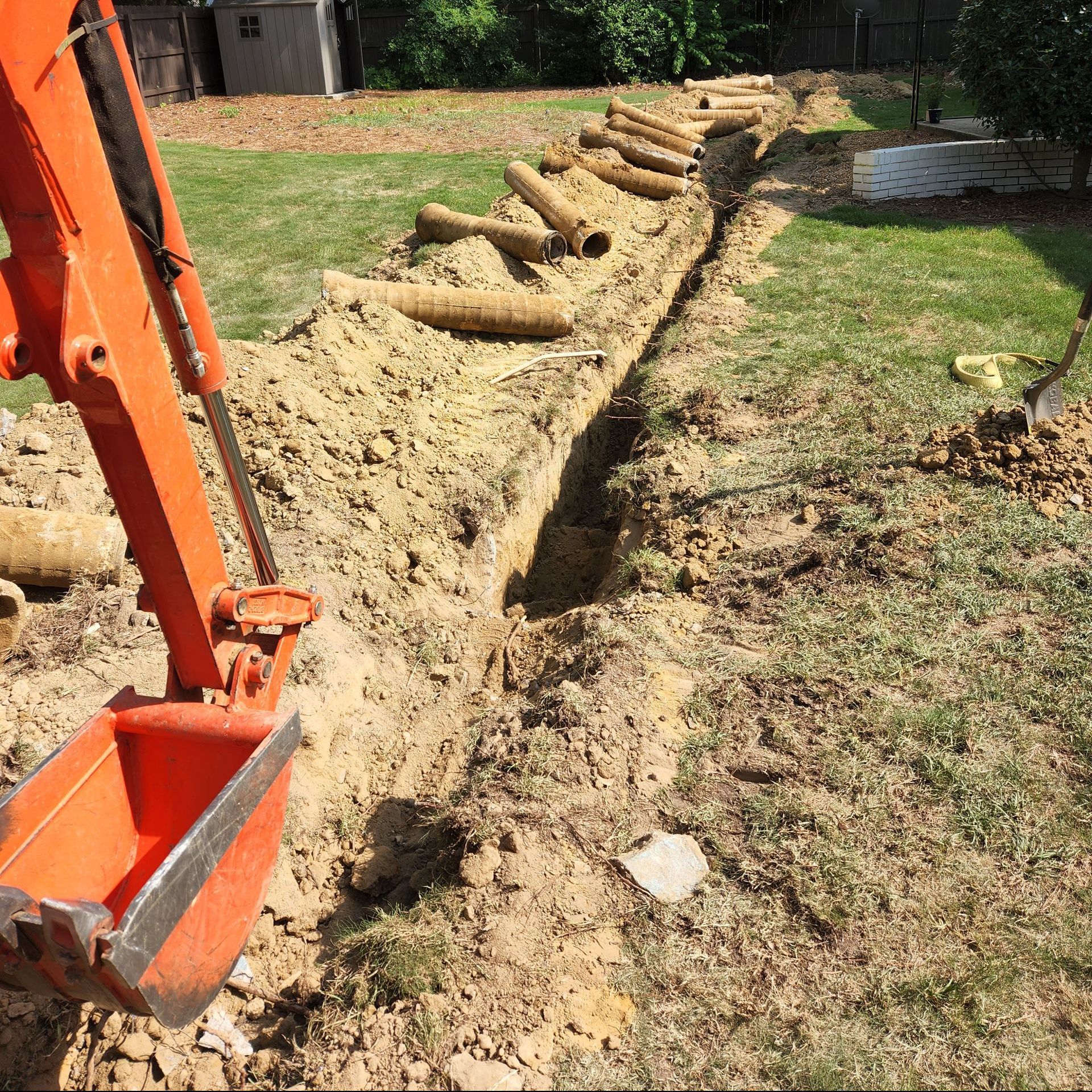An orange excavator bucket digs a long trench in a grassy yard, with pipes lined up nearby.
