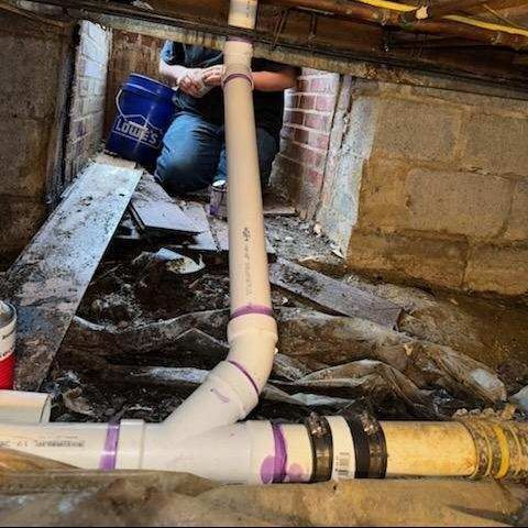 Plumber working on PVC pipes in a crawl space. A blue bucket is visible.