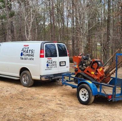 White Hays Plumbing van with trailer carrying a trencher in a wooded area.