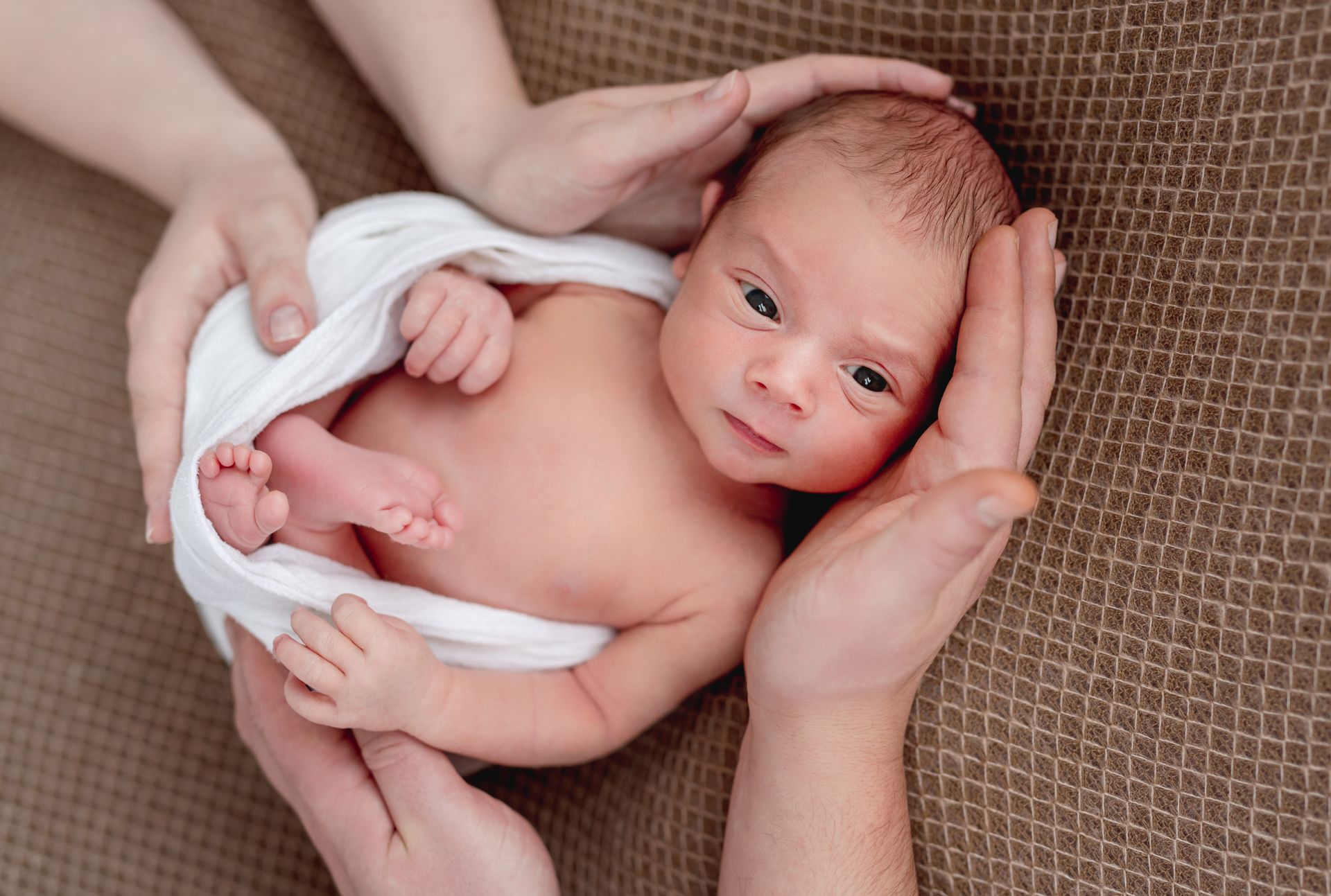 A newborn baby wrapped in a white blanket is being held by two people.