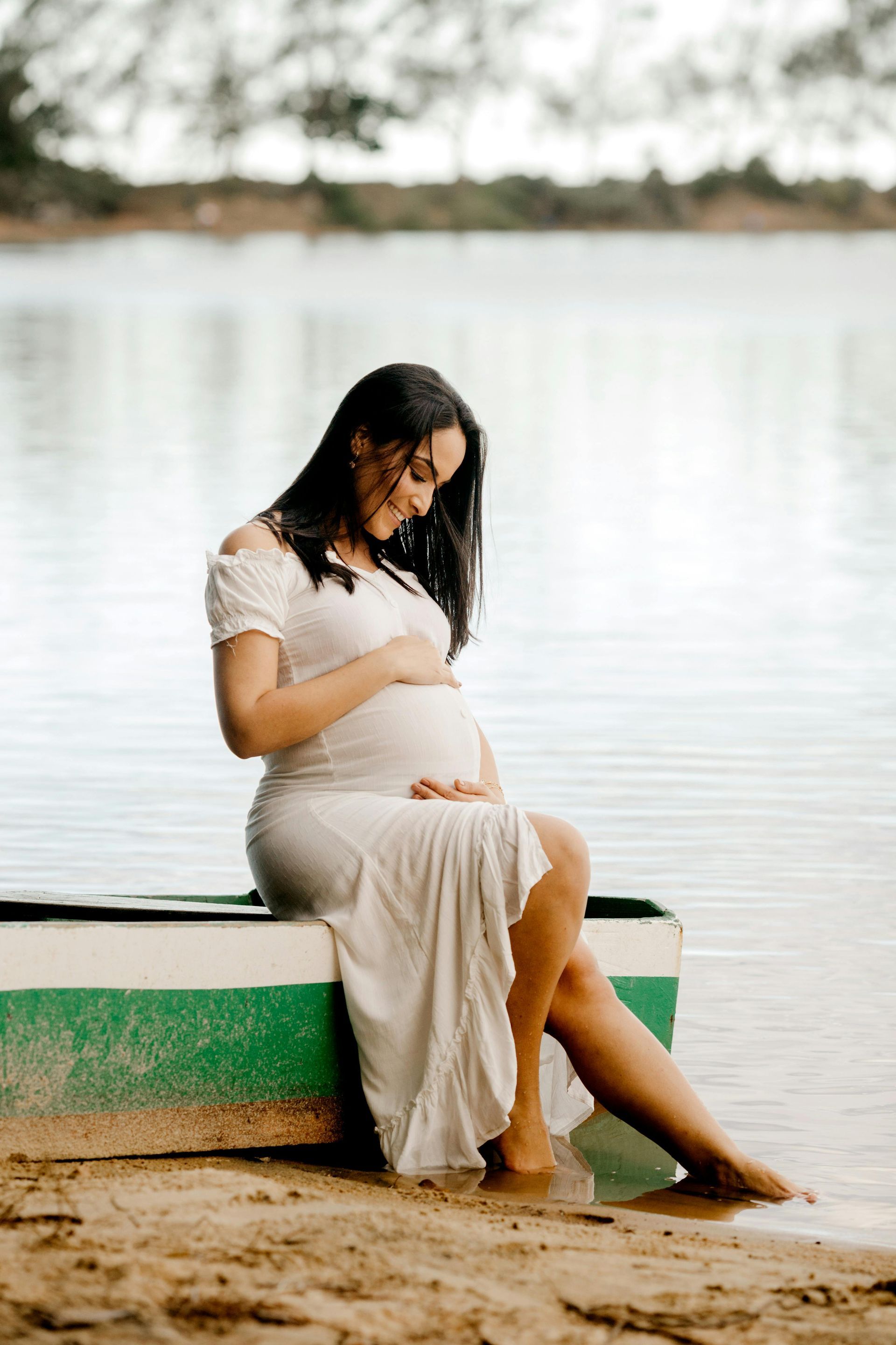 A pregnant woman is sitting on a boat on the beach holding her belly.
