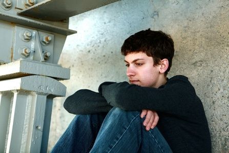 Young man in jeans and a dark sweater, sits leaning against a concrete wall, looking thoughtful.