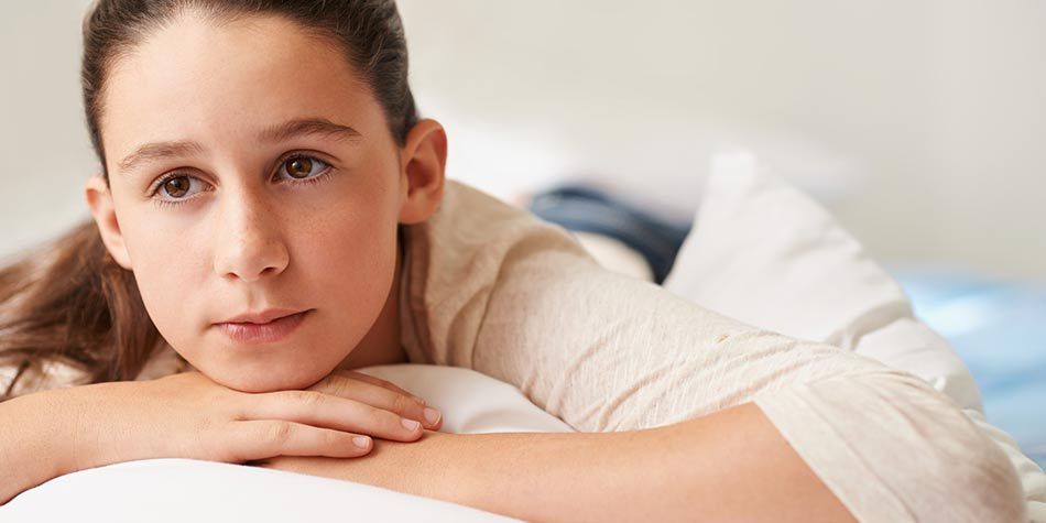 A girl with brown hair rests her head on her hands while lying on a bed, looking contemplative.