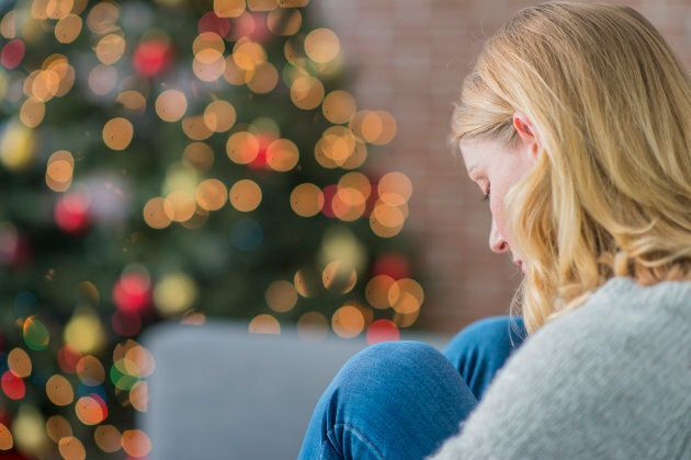 Woman with blonde hair looks down, curled up near a Christmas tree with bokeh lights.