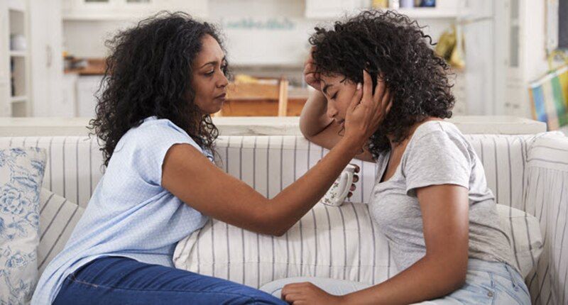 Mother comforts teenage daughter on a sofa; both appear upset in a home.