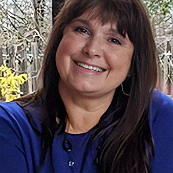 Woman with brown hair and bangs, smiling, wearing a blue shirt, outdoors with trees in the background.