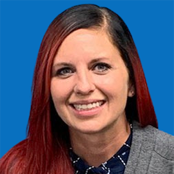 Woman with red hair smiling, wearing a gray cardigan over a patterned shirt, blue background.