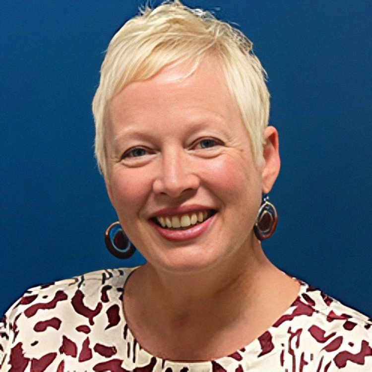 Woman with short blonde hair, smiling, wearing large hoop earrings and patterned blouse, against a blue background.