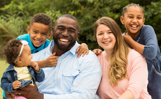 Family of five with diverse skin tones smiling outdoors.
