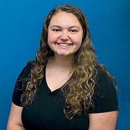 Woman with long curly brown hair and a smiling expression wearing a black shirt in front of a blue background.