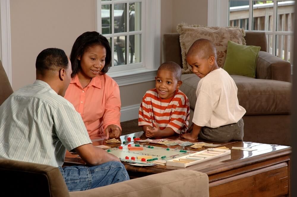 Family plays a board game in a living room. Parents and two young boys are seated around a table.