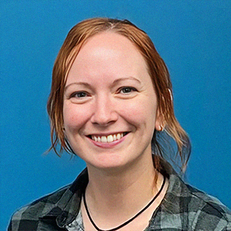 Woman with auburn hair, smiling, wearing a plaid shirt and necklace, blue background.