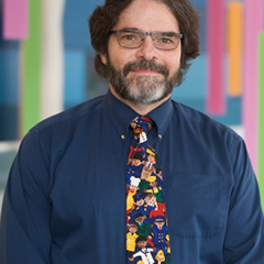 Man with glasses and beard wearing a blue shirt and colorful profession-themed tie, in a brightly colored hallway.
