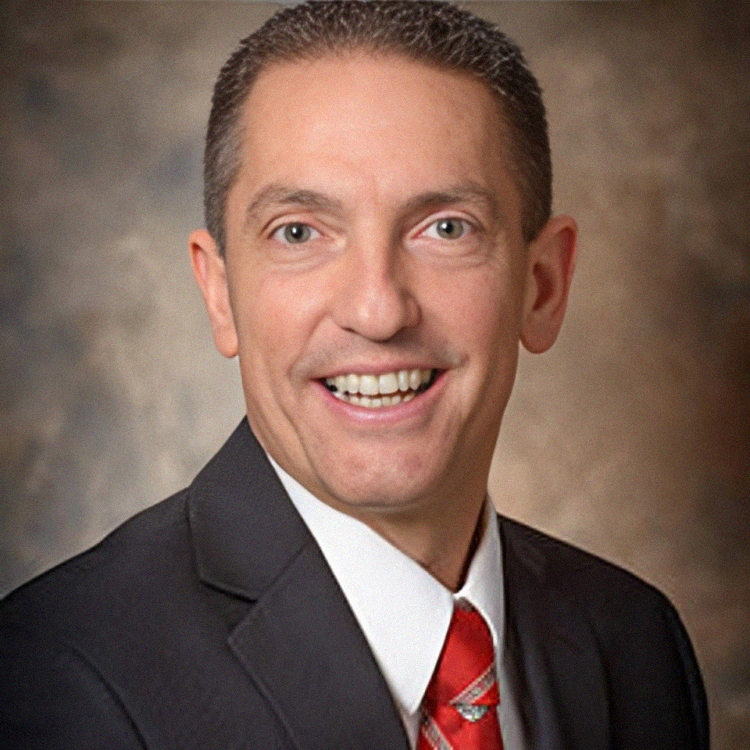 Man in suit smiles at camera, red tie, neutral background.