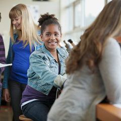 Girl in denim jacket smiles, sitting at a desk in classroom, two other students nearby.