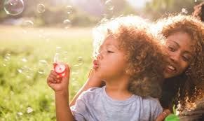 Child with curly hair blowing bubbles, smiling adult watches in a sunlit grassy area.