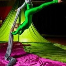 Person in a green bodysuit performing aerial silks, indoors. Silks are gray; fabric is pink and green.