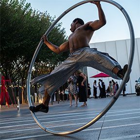 Man in silver pants performs acrobatic stunts inside a large metal wheel outdoors near a building.