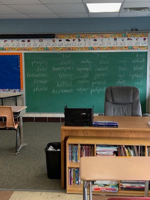 A classroom with a green chalkboard, desk, and bookshelves; a teacher's chair faces the board.
