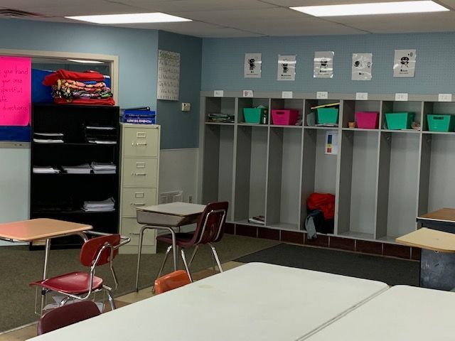 A classroom with desks, lockers, and a doorway. Blue walls, colorful bins, and student belongings.