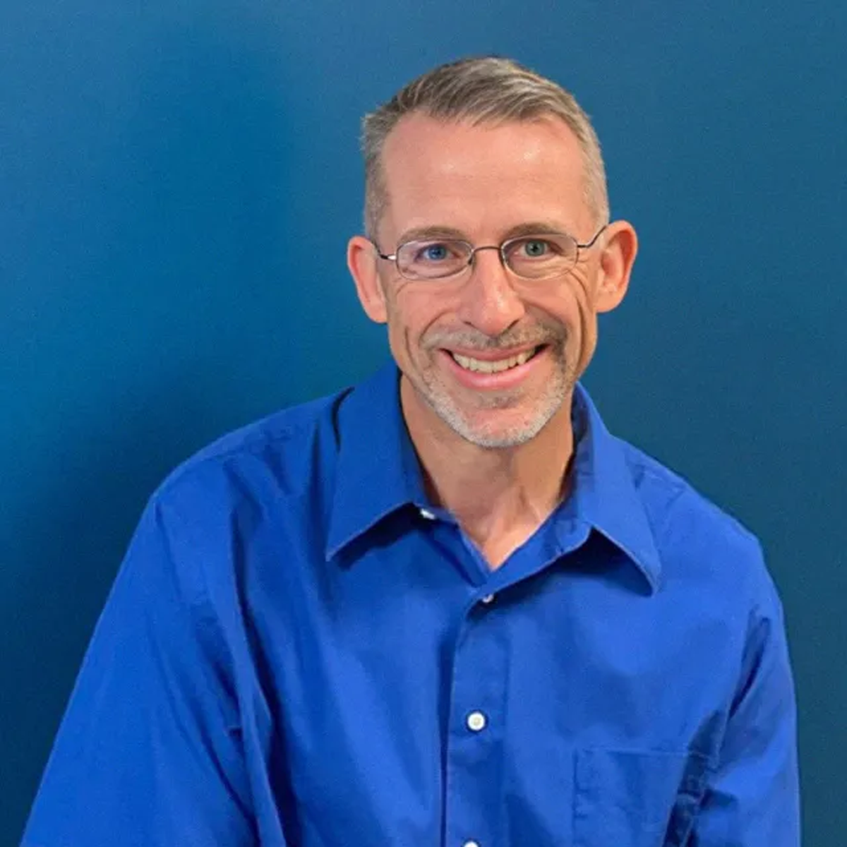 Man with graying hair and glasses, wearing a blue button-down shirt, smiles in front of a blue wall.
