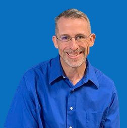 Smiling man with glasses in a blue shirt against a blue wall.