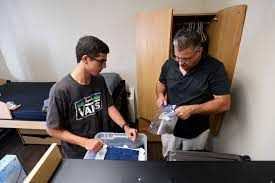 A young man and older man unpack belongings in a dorm room, near a wardrobe and a bed.