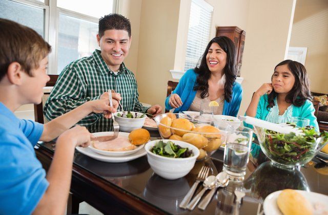Family of four eating at a dining table. They're smiling, eating salad and other foods. Bright interior setting.