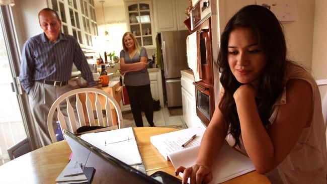 Teenage girl at table, using laptop. Parents watching in kitchen.