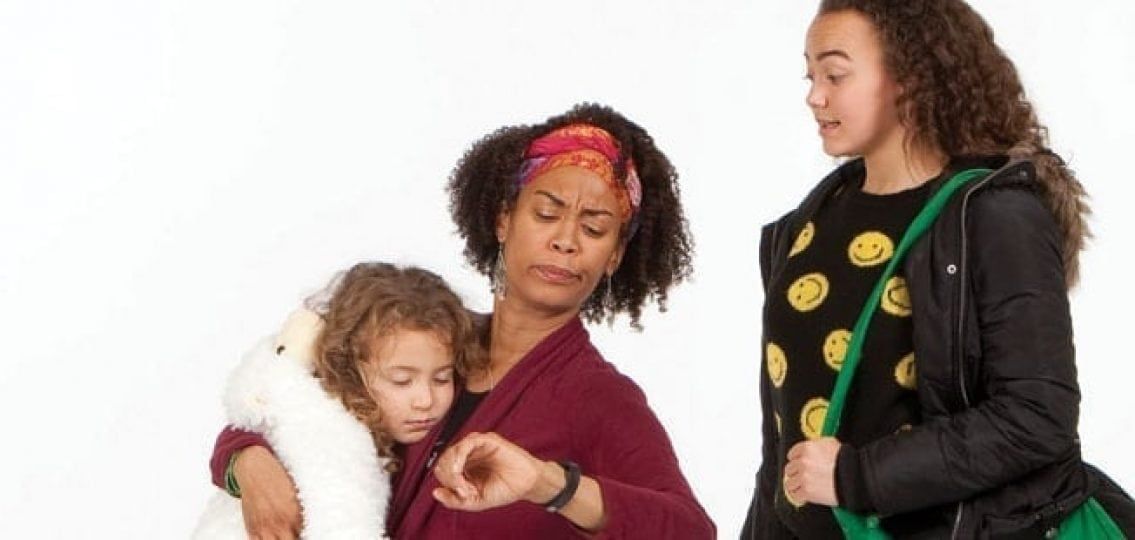 A mother checks her watch, holding a child. Teen daughter with a bag stands nearby. Studio white backdrop.