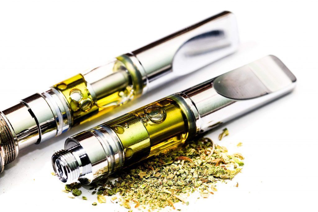 Two cannabis oil vape cartridges and dried cannabis buds on a white surface.